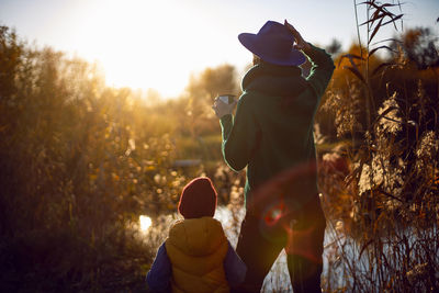 Woman in a hat and a boy stand with their backs to the lake at sunset in autumn