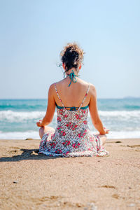 Rear view of woman meditating while sitting at beach against clear sky