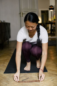 Young woman keeping incense stick on floor while crouching on exercise mat at home