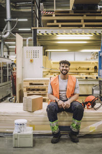 Portrait of happy young carpenter wearing reflective clothing sitting by hardhat in warehouse