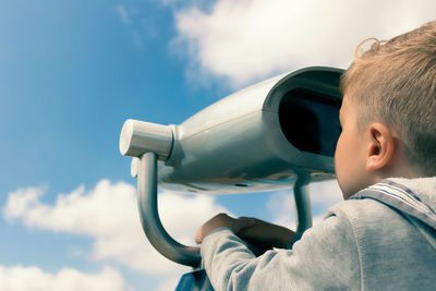 Little boy looking through coin operated binoculars against the sky.