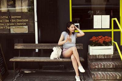 Thoughtful woman having coffee while sitting on bench at sidewalk cafe in city