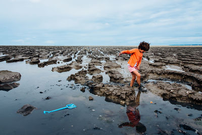 Side view of boy walking on rocky beach against sky