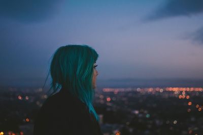 Side view of thoughtful woman standing against illuminated city at dusk