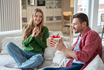 Overjoyed smiling woman giving gift present box to interested man congratulating sit on home couch