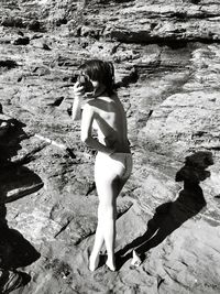 Shirtless young woman holding bikini top while standing at rocky beach during sunny day