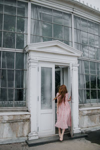 Full body of anonymous female in stylish outfit standing on street in doorway of hothouse with glass walls and growing green plants