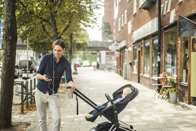 Smiling man using smart phone while standing by son on baby stroller in city