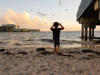 Rear view of boy standing on beach against sky during sunset