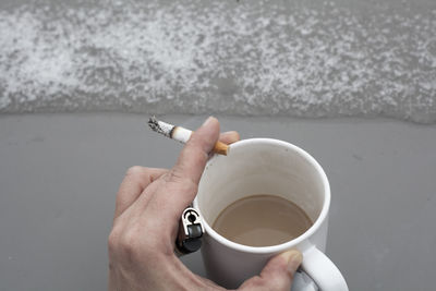 Cropped hand of man having coffee while smoking cigarette by wall