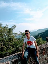 Young man sitting on retaining wall against sky