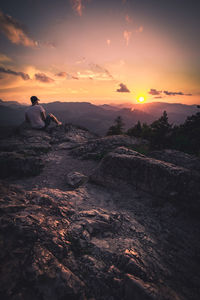 Rear view of man sitting on rock against sky during sunset