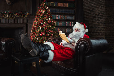 Full length of man wearing santa claus costume reading book on sofa at home