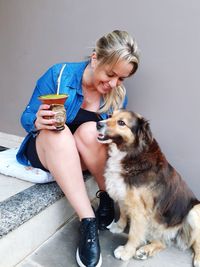 Full length of woman holding drink sitting with dog indoors