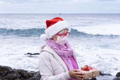 Senior woman wearing mask holding gift standing against sea