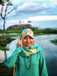 Portrait of smiling young woman showing peace sign against river