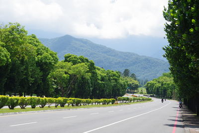 Empty road along trees and mountains against sky