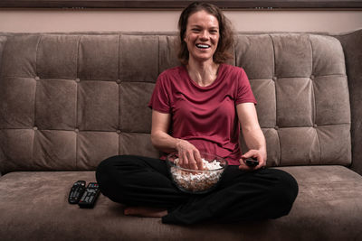 Full length of woman eating popcorn sitting on sofa at home