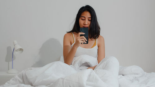 Young woman sitting on bed against wall at home