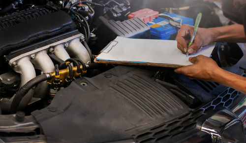 Cropped hands of mechanic writing on paper over car engine at garage