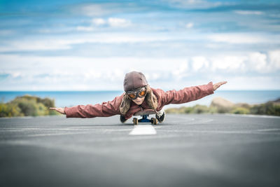 Portrait of boy lying on skateboard at street against cloudy sky