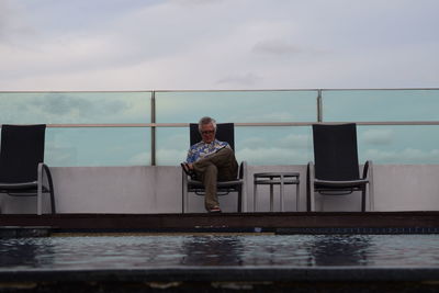 Senior man sitting on chair at poolside against cloudy sky