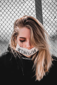 Young woman wearing face mask with text while sitting against chainlink fence