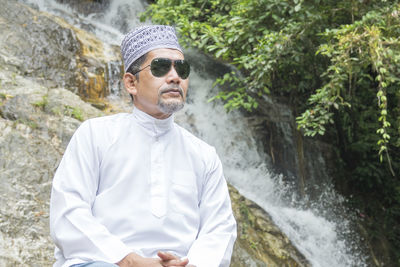 Mature man wearing sunglasses while sitting against waterfall