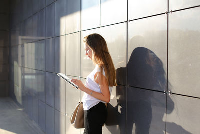 Young woman using digital tablet standing by wall