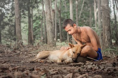 Shirtless teenage boy sitting with dog against trees in forest