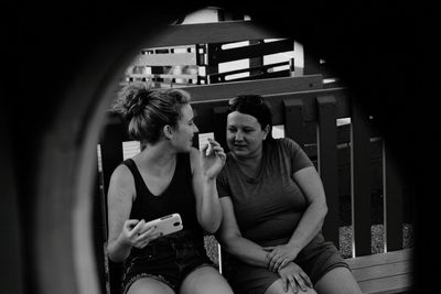 Female friends sitting on bench while using mobile phone at park seen through fence