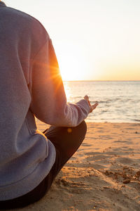 Midsection of man sitting at beach doing yoga on sunrise 