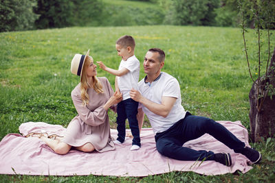 Family with a child a boy of 4 years old sitting on green field under a tree in summer on a blanket