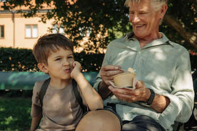 Boy leaning on elbow while sitting with grandfather holding ice cream cup at bench