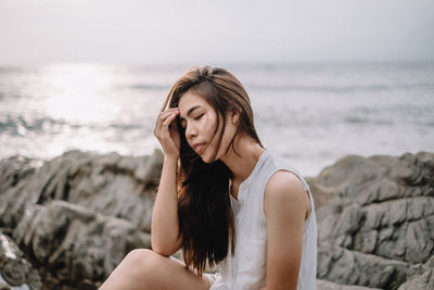 Beautiful young woman on rock at beach against sky