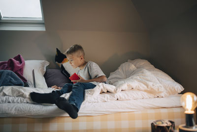 Full length of boy using laptop sitting with mobile phone on bed at home