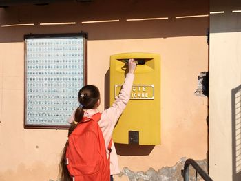Rear view of girl putting letter in mailbox on wall 