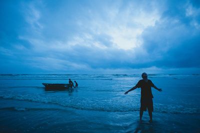 Man standing with arms outstretched on shore against cloudy sky