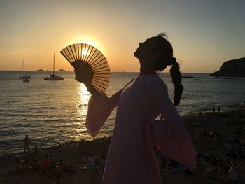 Woman holding folding fan against sea during sunset