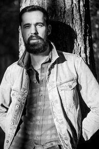 Portrait of bearded young man standing against tree trunk