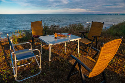 Empty chairs and table by sea against sky