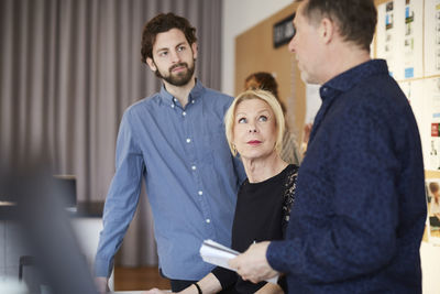 Senior businesswoman sitting amidst male colleagues against bulletin board at creative office