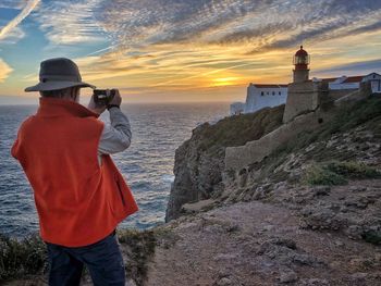 Rear view of man photographing lighthouse by sea against sky during sunset