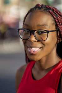 Positive african american female with red braids in eyeglasses showing tongue with piercing and looking at camera on street in sunny day