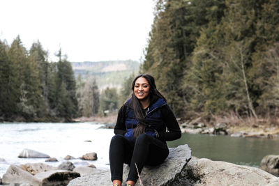 Portrait of smiling young woman sitting on rock by river