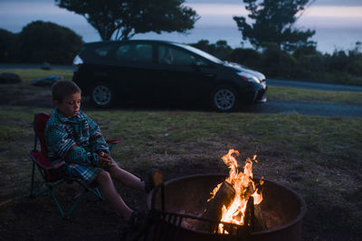 Full length of boy sitting on camping chair by campfire during dusk
