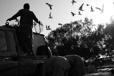 People with car against flock of birds in sky