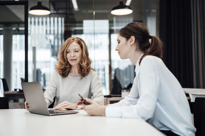 Female professionals discussing over laptop in illuminated board room at office