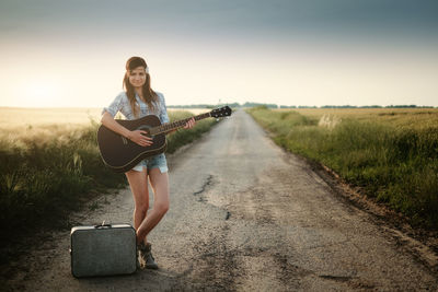 Portrait of smiling young woman playing guitar while standing on dirt road amidst field