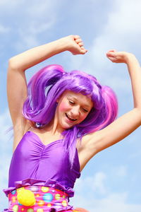 Low angle view of happy woman in costume and wig with arms raised against sky